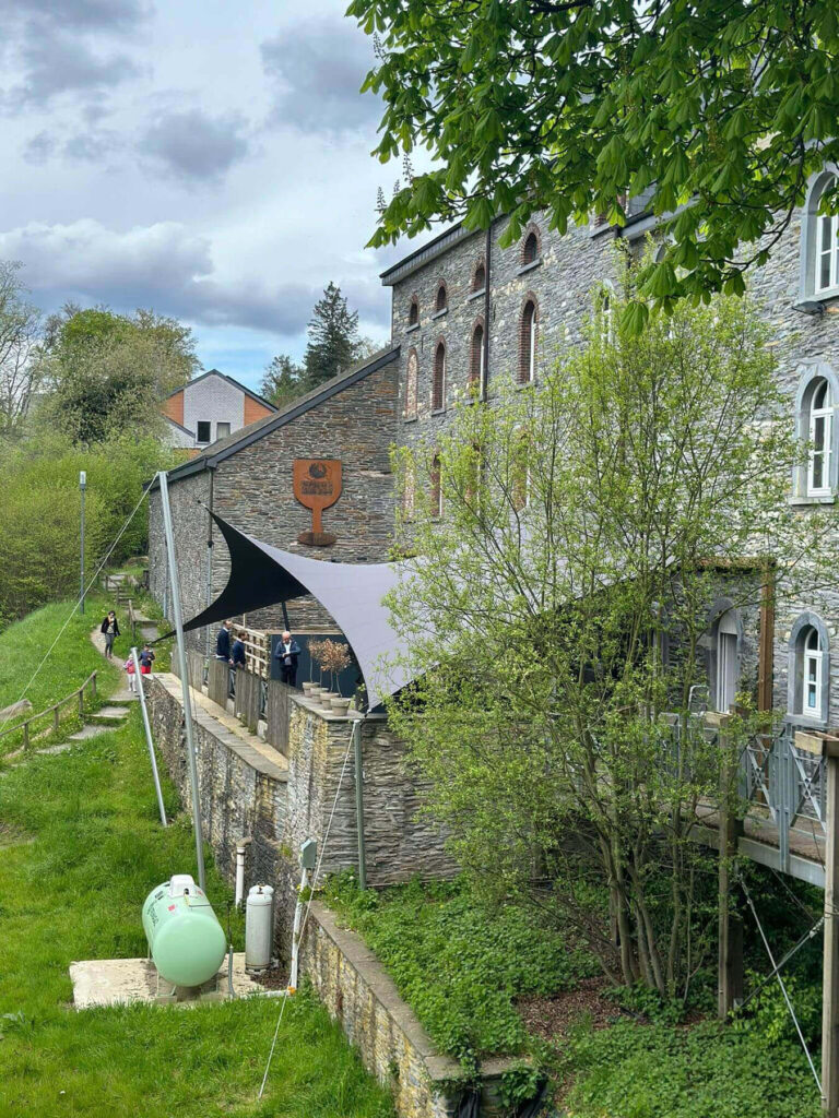 Stone building with arched windows beside a shaded outdoor terrace; people stand along a wooden railing under a large fabric canopy.