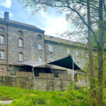 Stone building with arched windows and brick detailing, a wooden terrace with shade sails, and a grassy foreground.