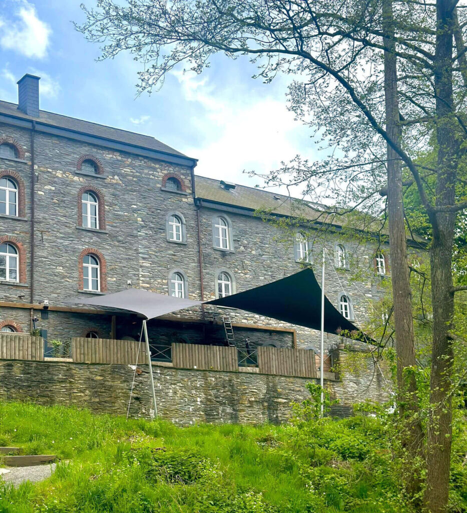 Stone building with arched windows and brick detailing, a wooden terrace with shade sails, and a grassy foreground.