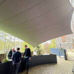 Group of three men standing and talking beside a long dark planter under a curved fabric shade sail in an outdoor area