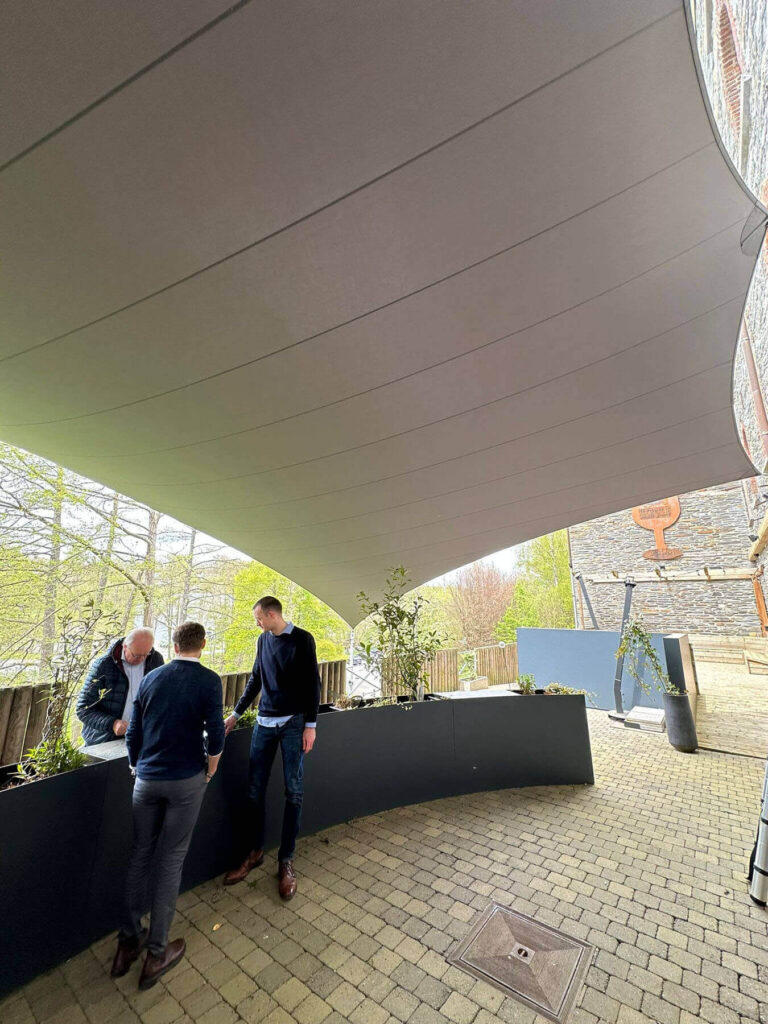 Group of three men standing and talking beside a long dark planter under a curved fabric shade sail in an outdoor area