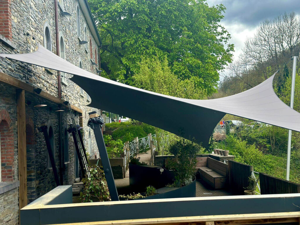 Outdoor patio beside a stone building with a large white sail-like shade canopy and lush green trees in the background.