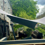 Outdoor patio beside a stone building with a large white sail-like shade canopy and lush green trees in the background.
