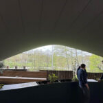 Interior of a curved-ceiling pavilion with dark walls, wooden planters, and a view of trees outside; two people on the right side.