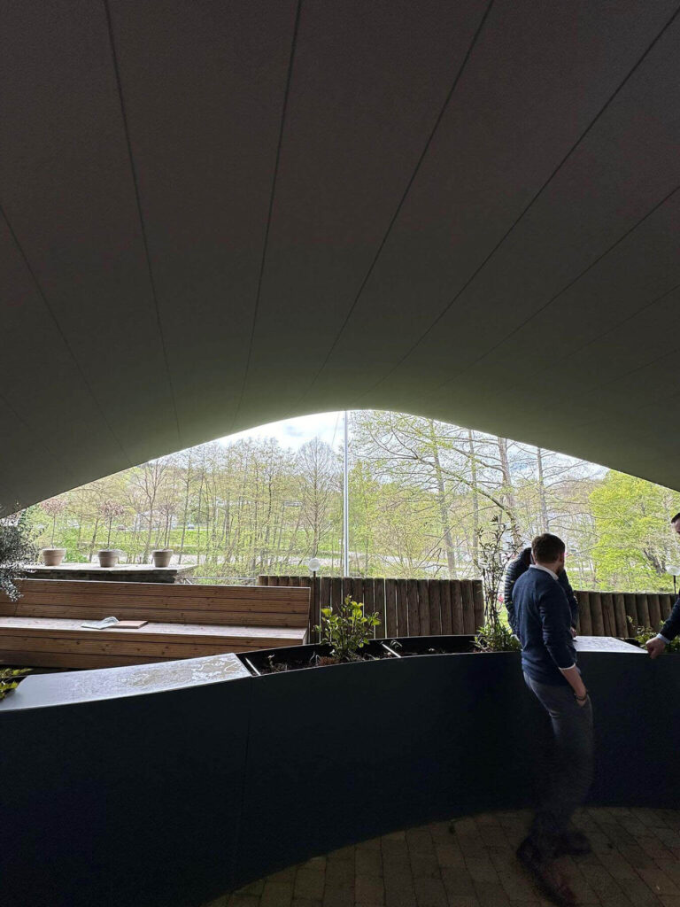Interior of a curved-ceiling pavilion with dark walls, wooden planters, and a view of trees outside; two people on the right side.