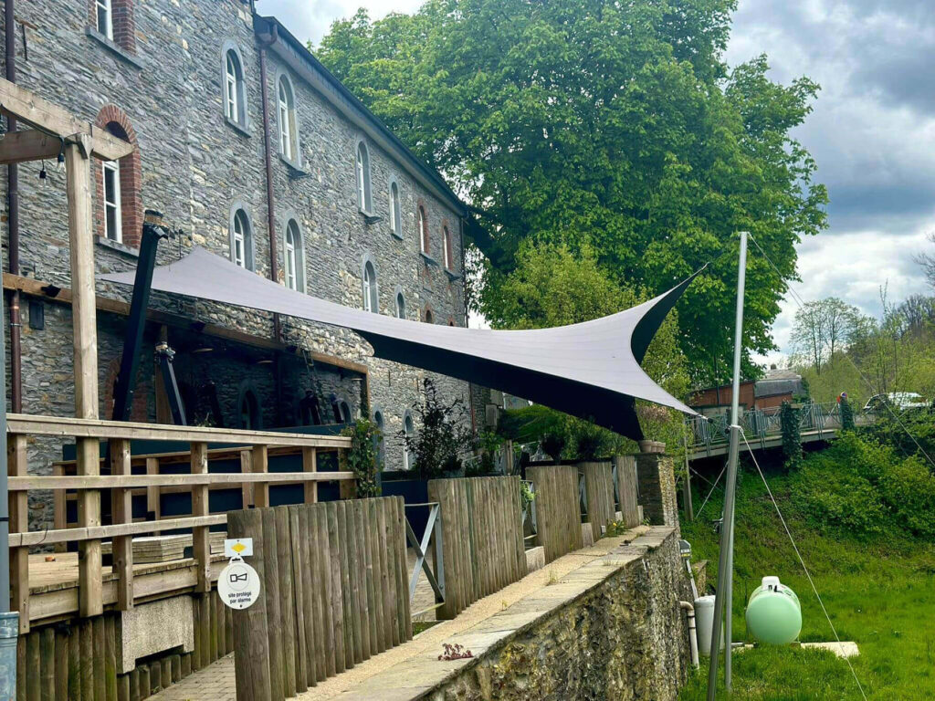 Stone building with arched windows and a large fabric shade sail over a wooden terrace, green trees in the background and a cloudy sky.