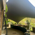 Outdoor patio with a large gray shade sail stretched between a brick wall and posts over a brick walkway, with potted plants along the border