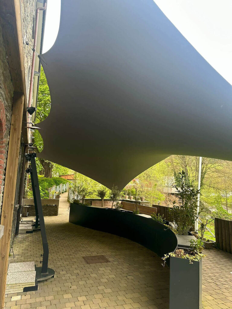 Outdoor patio with a large gray shade sail stretched between a brick wall and posts over a brick walkway, with potted plants along the border