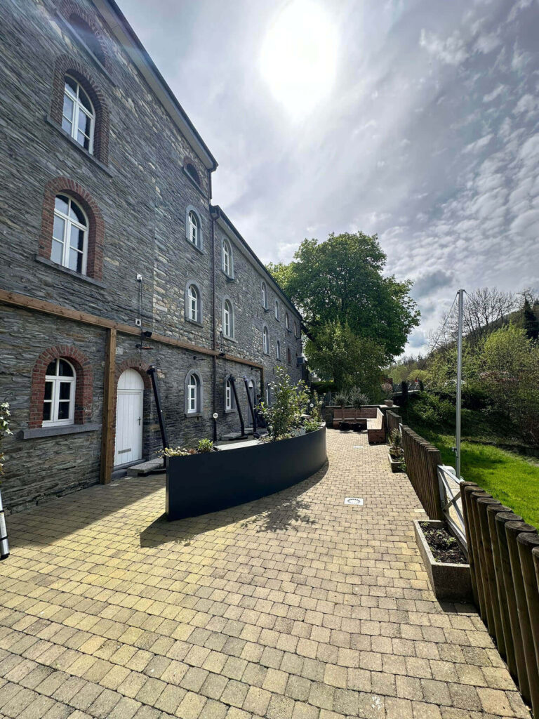Stone building with red-brick arched window frames along a paved courtyard and a wooden fence on the right.