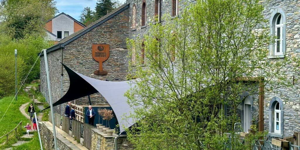 Stone building with arched windows and a curved grey shade sail over a terrace where a few people stand along a stone railing. A grassy path and trees run beside the scene.