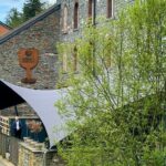 Stone building with arched windows and a curved grey shade sail over a terrace where a few people stand along a stone railing. A grassy path and trees run beside the scene.