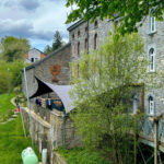 Stone historic building with arched windows and a curved terrace, where visitors stand along a stone walkway under a large shade sail on a sunny day.