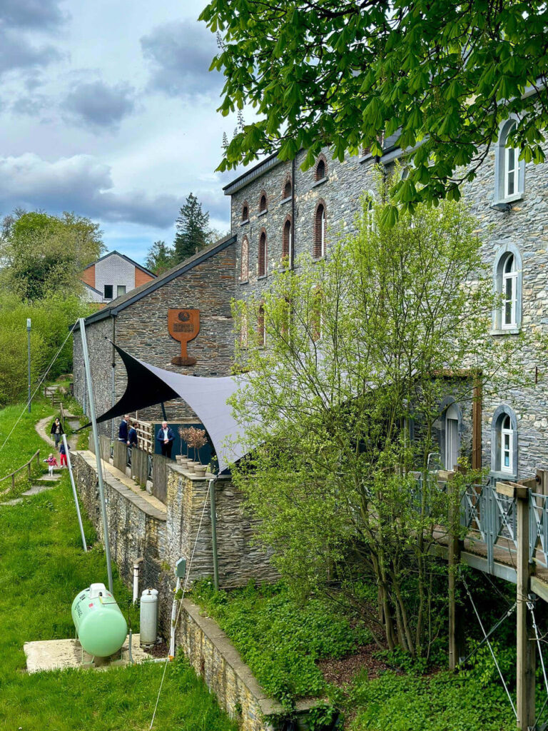 Stone historic building with arched windows and a curved terrace, where visitors stand along a stone walkway under a large shade sail on a sunny day.