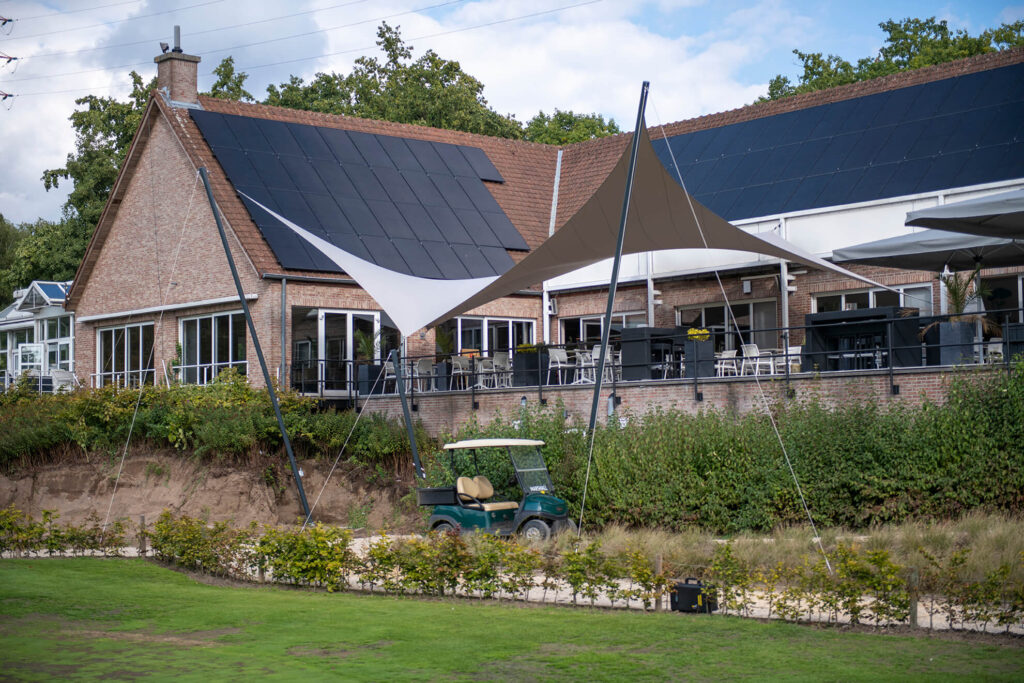 A brick building with dark solar panels on the sloped roof, outdoor seating under tan and white shade sails, and a green golf cart in the foreground.
