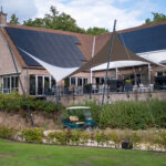A brick building with dark solar panels on the sloped roof, outdoor seating under tan and white shade sails, and a green golf cart in the foreground.