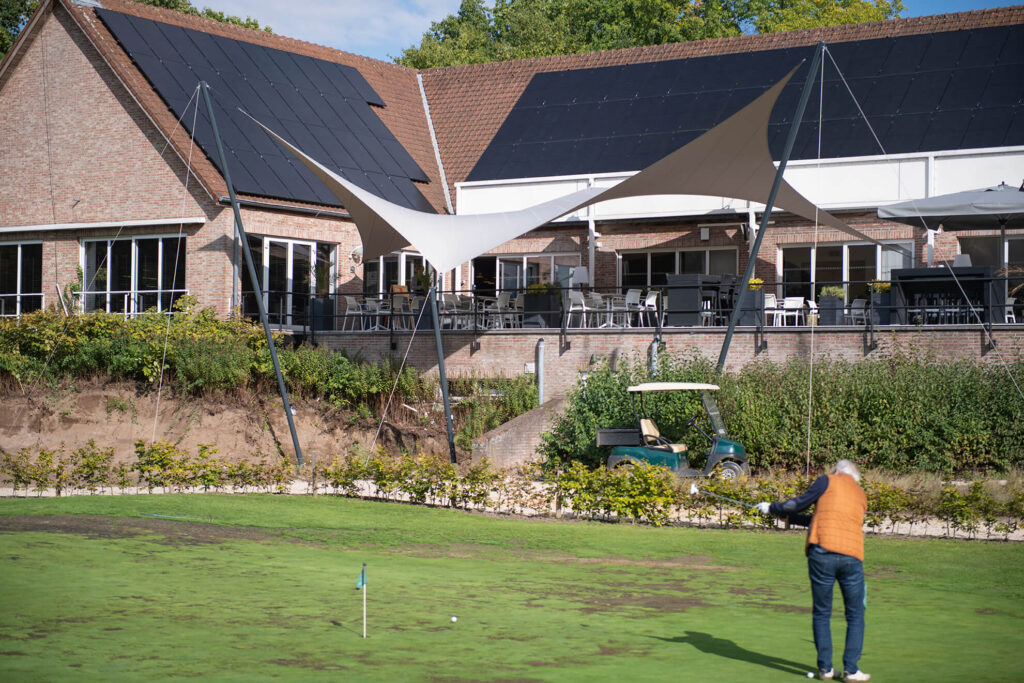 Man in an orange jacket stands on a grassy putting area near a brick building with solar panels and a shaded outdoor seating area.