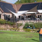 Man in an orange jacket stands on a grassy putting area near a brick building with solar panels and a shaded outdoor seating area.