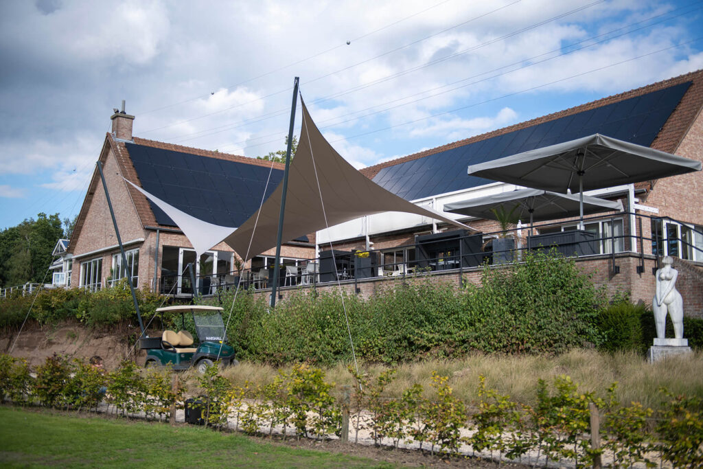 Brick building with solar panels on the sloped roof, beige shade sails over an outdoor seating area, and a statue in the foreground right.