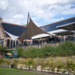 Brick building with solar panels on the sloped roof, beige shade sails over an outdoor seating area, and a statue in the foreground right.