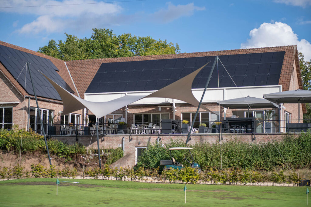 Building with large solar panels on the roof, outdoor seating under white and beige shade sails, and a green lawn in front.