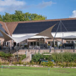 Building with large solar panels on the roof, outdoor seating under white and beige shade sails, and a green lawn in front.