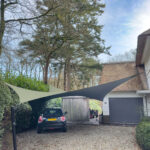 Residential driveway with a dark car parked under two triangular shade sails, tall trees and a house with a gray garage door in the background.