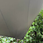 Gray outdoor umbrella canopy with stitched seams, set against green shrubbery.