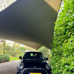 Black Mini Cooper parked in a gravel driveway under a large angled shade sail with green hedges on the right.