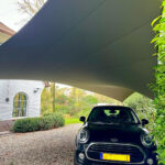Black Mini Cooper parked under a large grey shade sail in a residential gravel driveway, beside a white brick house with a window.