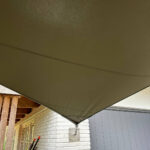 Brown shade sail stretched over a brick patio, attached to a wooden pergola and exterior wall.