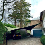 Driveway scene with gravel, a black car parked under two fabric shade sails mounted on poles beside a house with a blue garage door.