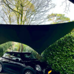 Black Mini Cooper parked on a gravel driveway under a large curved black shade sail, with trees and hedges in the background.