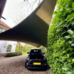 Black compact car parked on a gravel driveway under a curved fabric shade sail, with a tall green hedge on the right and a house in the background