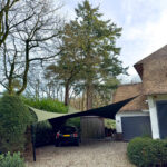 Gravel driveway with a black car parked under two large tan and dark sails, beside a thatched-roof house and tall trees in the yard.