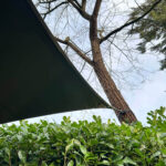 Dark green triangular shade sail attached to a tree trunk, with a leafy hedge in the foreground and blue sky.