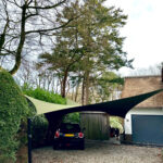 SUV parked under green and dark shade sails in a gravel driveway beside a hedge and a house with a blue garage door