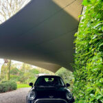 Black Mini Cooper parked in a gravel driveway under a large gray shade sail, with a green hedge on the right.