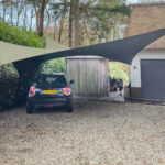 Black Mini Cooper parked under beige and dark gray shade sails in a gravel driveway beside a white house with a gray garage door; trees in the background.