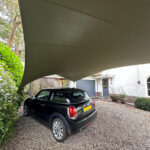 Black Mini Cooper parked on a gravel driveway under a large curved beige shade sail in front of a white house with greenery nearby.