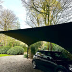 Black SUV parked on a gravel driveway beneath a curved dark shade sail, with trees and hedges in the background.