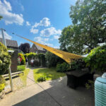 Backyard with a large yellow shade sail over an outdoor table; two people stand near a brick path under a blue sky.