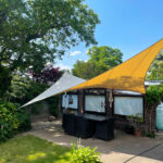 Outdoor patio with a dining table and black chairs under a large white and yellow sail shade in a sunny backyard with trees and brick house facade
