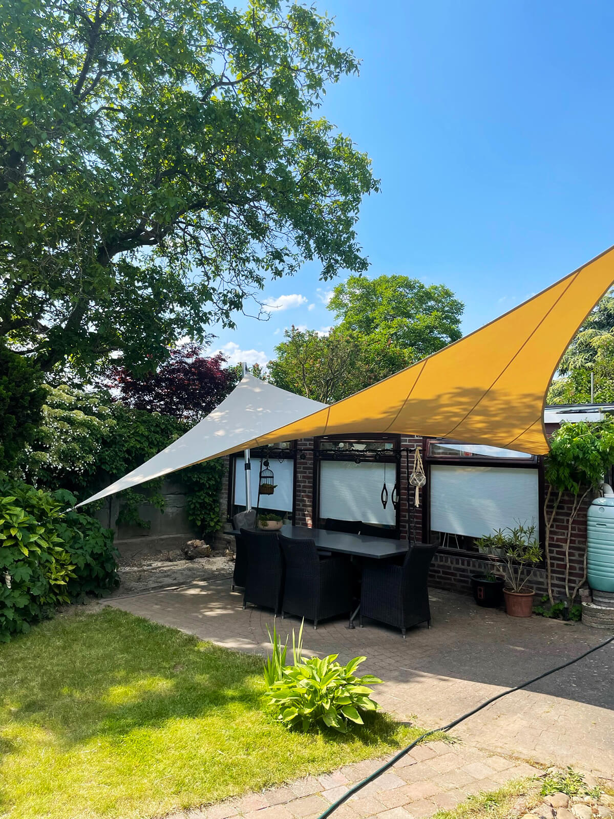 project-Stein-tuindoek-op-maat-terras-overkapping-zonnedoek-key-doek-stormvast-verwijderbaar_02 Outdoor patio with a dining table and black chairs under a large white and yellow sail shade in a sunny backyard with trees and brick house facade