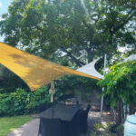 Outdoor patio dining area beneath a large tree, with yellow and white shade sails and black wicker chairs nearby.