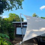 Large white fabric shade sail stretched over a paved backyard area, supported by a metal pole beside a brick house with green plants and trees nearby.
