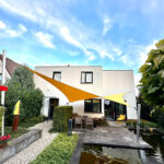 Modern white house with orange shade sails over a stone patio and dining area, under a blue sky.