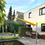 Outdoor patio with a rectangular pool, tan paving, and a yellow-orange shade sail stretched between posts above a dining table and chairs by the house.