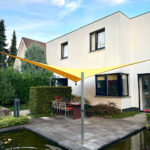 Modern white two-story house with bright yellow shade sails over a stone patio dining area by a rectangular pond, sunny day.