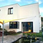 Modern two-story white house with large glass doors opening to a patio, yellow awning, and outdoor seating overlooking a rectangular shallow pond with lily pads and a duck statue.