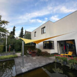 Backyard patio of a modern white house with a yellow shade sail over a dining table and yellow chairs beside a rectangular pond.